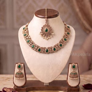 May include: A jewelry set featuring a necklace, a pair of jhumka earrings, and a maang tikka. The pieces are crafted in a gold-tone metal with intricate filigree detailing, set with teardrop-shaped green stones and small white faux pearls.