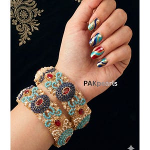May include: Two ornate gold bangles with turquoise, red, and navy blue accents. The bangles are adorned with small pearls and gemstones. The image also shows a hand with matching nail art. The text "PAKpearls" is visible.