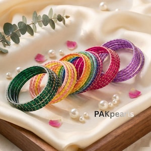 May include: A selection of vibrant bangles in green, yellow, pink, red, and purple. The bangles are displayed on a cream satin surface, decorated with pearls and rose petals. These bangles are a lovely accessory.