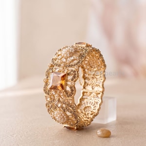 May include: Ornate gold-toned bracelet with a large, square amber-colored gemstone at its center. The bracelet features intricate filigree and is adorned with small, pearl-like beads and clear crystals. A small, oval-shaped stone sits near the bracelet.