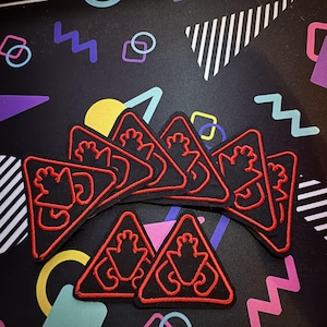 May include: A collection of black triangular patches with a red border and a stylized red graphic. The patches are arranged on a black surface with colorful geometric shapes. The patches are likely for clothing or accessories.
