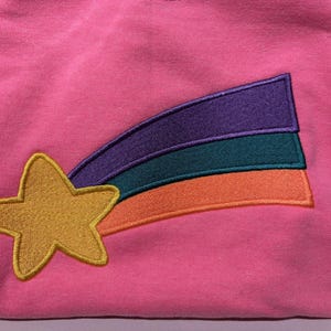 May include: Pink sweatshirt featuring an embroidered design of a yellow star and a shooting star. The shooting star has purple, teal, and orange stripes. The design is on the front of the garment.