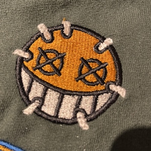 Op de afbeelding: Geborduurde patch met een lachend gezicht met gestikte details. Het gezicht heeft een gele kern, zwarte doorgestreepte ogen en een witgetande grijns omlijnd in zwart. De patch is op een groene stof genaaid.