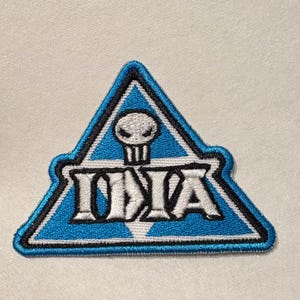 May include: Embroidered patch in a triangular shape with a light blue background and black border. The patch features the word "IDIA" in white block letters, with a skull icon above the letters.