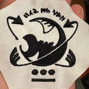 May include: A white fabric patch with a black embroidered design. The design features a stylized creature with curved lines, arrows, and Japanese text. Below the creature are three dots and a horizontal line.