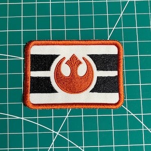 Può includere: Toppa rettangolare ricamata con sfondo bianco, strisce bianche e nere e bordo arancione. La toppa presenta un emblema arancione al centro. Probabilmente per abbigliamento o accessori.