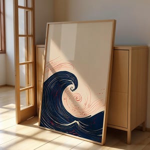 May include: A framed art print featuring a stylized wave design. The artwork showcases a large, dark blue wave with white accents and red swirling lines against a cream background. The frame is a light wood color, and the print leans against a wooden cabinet.