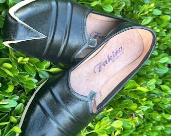 May include: A pair of black leather slip-on shoes with a pointed toe and a white trim. The shoes have a textured design and a light brown interior. The shoes are placed on a bed of green leaves.