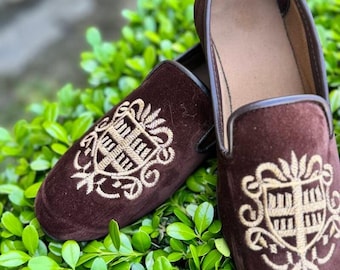 Op de afbeelding: Een paar bruine fluwelen loafers met een geborduurd gouden wapenontwerp. De schoenen hebben een klassieke instapstijl en staan tegen een achtergrond van groen gebladerte. De loafers zijn een rijke, donkerbruine kleur.