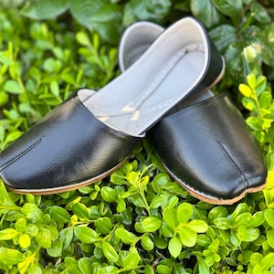 Puede incluir: Un par de bailarinas de cuero negro con punta redondeada y suela fina de color claro. Los zapatos se exhiben sobre un lecho de follaje verde, mostrando su diseño y textura. Las bailarinas tienen un diseño simple y clásico.