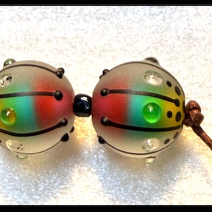 May include: Two spherical glass beads strung on a brown cord, featuring a frosted finish with a rainbow-colored gradient of red, green, and yellow. The beads are decorated with black lines, small black dots, and embedded clear glass accents.