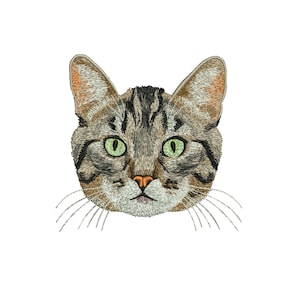 Pode incluir: Rosto de gato bordado com olhos verdes, nariz rosa e bigodes. A pelagem do gato é uma mistura de castanho, cinza e preto, com costuras detalhadas. A cabeça do gato está centrada em um fundo branco.