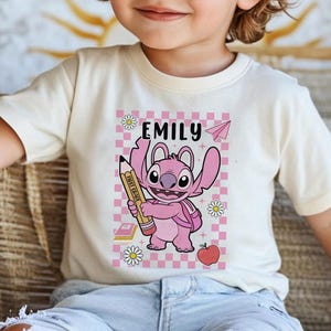 以下が含まれることがあります： ピンク色の漫画のキャラクターが鉛筆を持っているクリーム色のTシャツ。Tシャツには、キャラクターの上に「Emily」という名前があり、デイジー、紙飛行機、本、リンゴのあるピンクと白のチェック柄の背景が描かれています。