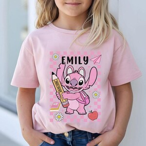 以下が含まれることがあります： 鉛筆を持った漫画のキャラクターが描かれたピンク色のTシャツ。「EMILY」という名前がキャラクターの上にあり、紙飛行機とデイジーのアクセントが付いています。キャラクターはピンク色で、大きな目とバックパックが付いています。