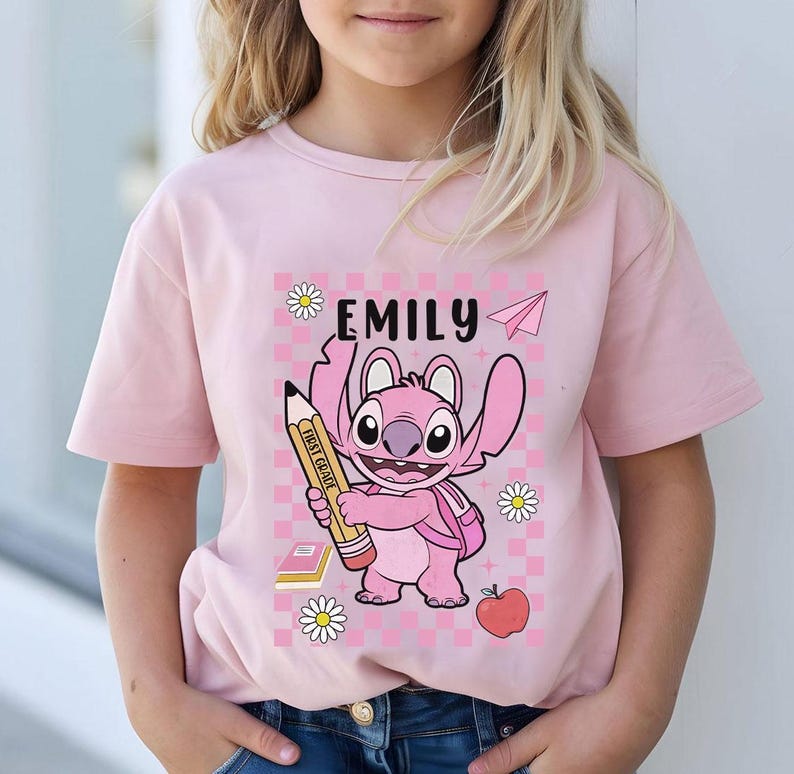 以下が含まれることがあります： 鉛筆を持った漫画のキャラクターが描かれたピンク色のTシャツ。「EMILY」という名前がキャラクターの上にあり、紙飛行機とデイジーのアクセントが付いています。キャラクターはピンク色で、大きな目とバックパックが付いています。