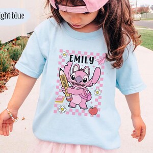 以下が含まれることがあります： ピンクの漫画のキャラクターが鉛筆を持っているライトブルーのTシャツ。上部に「EMILY」という名前があります。デザインには、ピンクと白のチェック柄の背景、紙飛行機、デイジー、本、リンゴが含まれています。