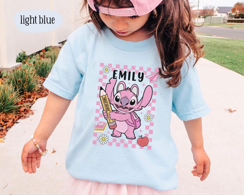 以下が含まれることがあります： ピンクの漫画のキャラクターが鉛筆を持っているライトブルーのTシャツ。上部に「EMILY」という名前があります。デザインには、ピンクと白のチェック柄の背景、紙飛行機、デイジー、本、リンゴが含まれています。