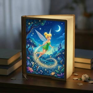 May include: A book-shaped lamp with a fairy illustration, emitting a warm glow. The image shows a fairy in a green dress, with a trail of light, a moon, and a castle. The lamp is on a wooden surface, creating a magical atmosphere.