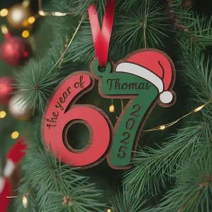 May include: A personalized Christmas ornament with a red and green color scheme. The ornament features the number "67" in red, the text "the year of" and "2025" in green, and a Santa hat. The name "Thomas" is also displayed. A red ribbon is attached for hanging.