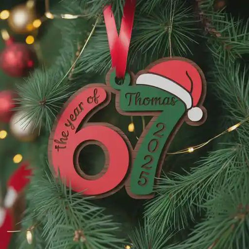 May include: A personalized Christmas ornament with a red and green color scheme. The ornament features the number "67" in red, the text "the year of" and "2025" in green, and a Santa hat. The name "Thomas" is also displayed. A red ribbon is attached for hanging.