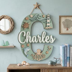 May include: A large, sage green wooden letter C wall hanging featuring the name Charles in white script. The letter is decorated with layered nautical elements including a lighthouse, a sea turtle, a dolphin, an octopus, coral, and seashells.