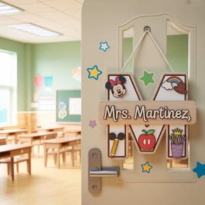 May include: Wooden door sign in the shape of the letter "M" with the name "Mrs. Martinez". The sign features cartoon images of Minnie Mouse, a rainbow, an apple, and pencils. The sign is hanging on a door in a classroom.