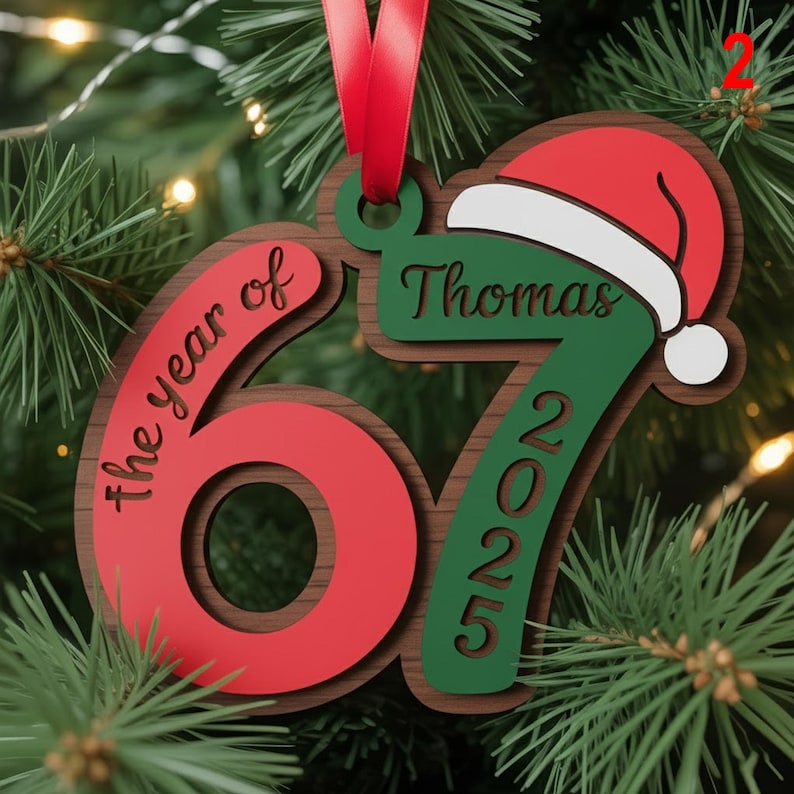 May include: A wooden Christmas ornament with a red and green design. The ornament features the number "67" in red, the words "the year of" and "2025". A Santa hat and the name "Thomas" are also included.