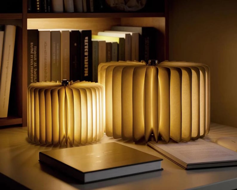 May include: Two illuminated paper lamps, resembling open books, sit on a desk. The lamps are made of folded paper, emitting a warm, inviting glow. A closed book and an open book are on the desk. Bookshelves are in the background.