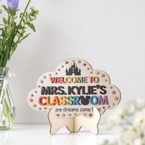 May include: A wooden classroom sign in the shape of a cloud, with the words "WELCOME TO MRS. KYLIE'S CLASSROOM" in colorful letters. The sign features a castle silhouette and star accents. It stands on a wooden base.