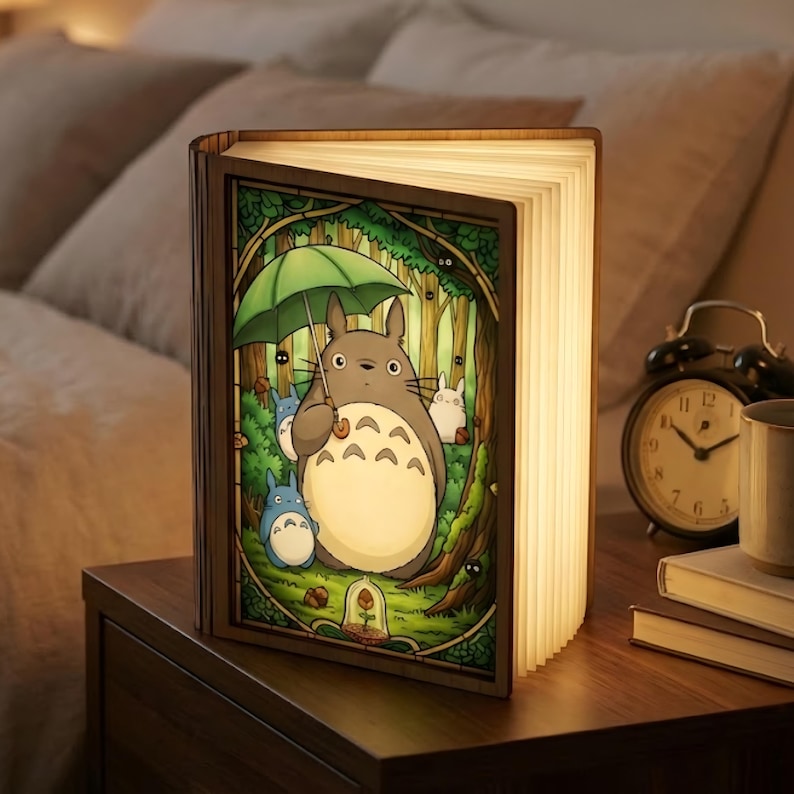May include: A wooden book-shaped lamp featuring a colorful illustration of Totoro and other characters from the animated film. The lamp is open, with the pages illuminated, and sits on a wooden nightstand.