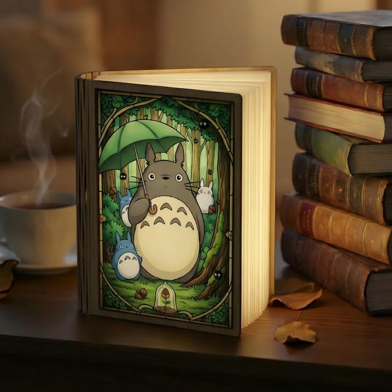 May include: A book-shaped lamp with a colorful illustration of Totoro and other characters from the animated film. The lamp is open, with the pages illuminated, and is sitting on a wooden surface. A stack of books is in the background.