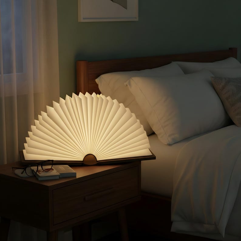 May include: A unique, open-book-shaped LED lamp with a warm, inviting glow. The lamp sits on a wooden nightstand next to a bed with white bedding. The lamp's pages are fanned out, creating a soft, ambient light source for a bedroom.