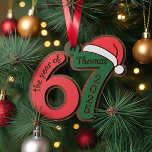 May include: A wooden Christmas ornament with the text "the year of 67 2025" and the name "Thomas" with a Santa hat. The ornament is red, green, and brown, with a red ribbon for hanging. The ornament is hanging on a Christmas tree.
