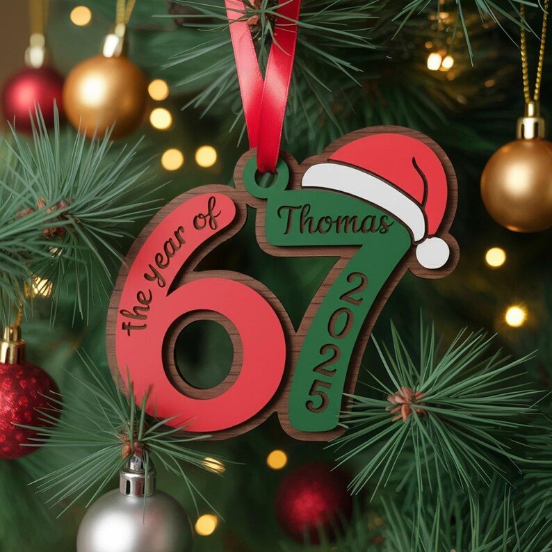 May include: A wooden Christmas ornament with the text "the year of 67 2025" and the name "Thomas" with a Santa hat. The ornament is red, green, and brown, with a red ribbon for hanging. The ornament is hanging on a Christmas tree.
