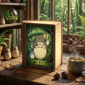 May include: A wooden book-shaped lamp featuring a colorful illustration of Totoro characters in a forest scene. The lamp is open, emitting a warm glow, and sits on a wooden surface with acorns and a cup of tea. Other Totoro figurines are nearby.