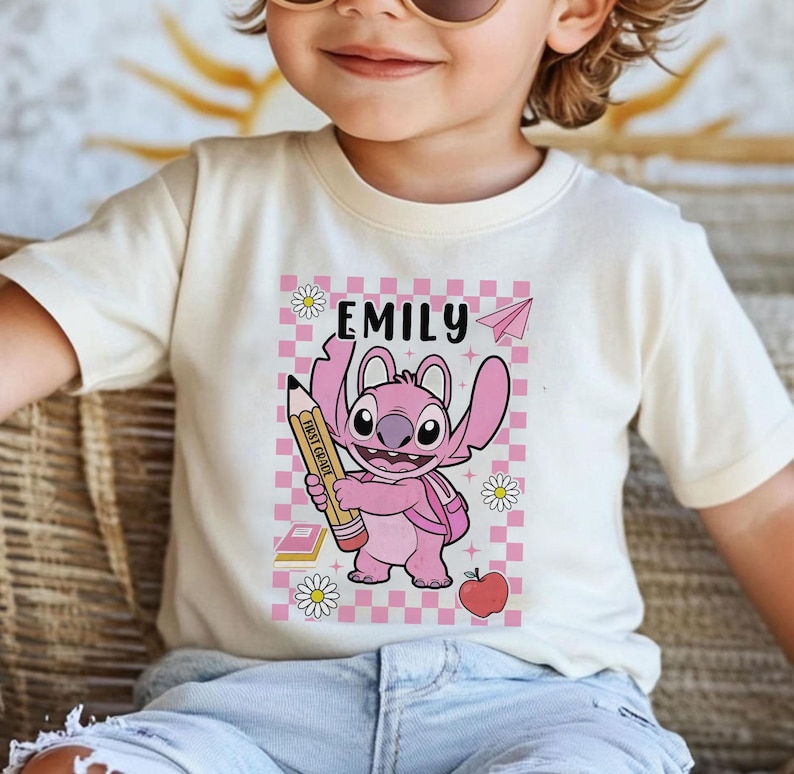 以下が含まれることがあります： ピンク色の漫画のキャラクターが鉛筆を持っているクリーム色のTシャツ。Tシャツには、キャラクターの上に「Emily」という名前があり、デイジー、紙飛行機、本、リンゴのあるピンクと白のチェック柄の背景が描かれています。