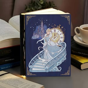 May include: A navy blue, wooden book-shaped lamp with a Cinderella theme. The cover features Cinderella in a light blue gown, a clock, and a castle. The lamp is open, revealing a warm glow, and is surrounded by other books.