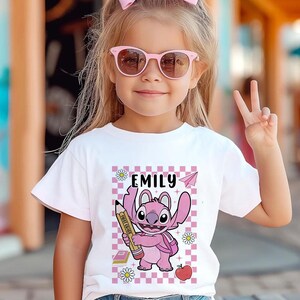 以下が含まれることがあります： 白いTシャツには、鉛筆を持ったピンク色の漫画のキャラクターが描かれており、その上に「EMILY」という名前があります。 デザインには、ピンクと白のチェック柄の背景、紙飛行機、本、デイジー、リンゴが含まれています。