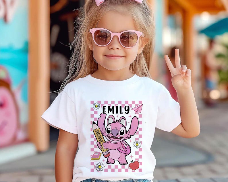 以下が含まれることがあります： 白いTシャツには、鉛筆を持ったピンク色の漫画のキャラクターが描かれており、その上に「EMILY」という名前があります。 デザインには、ピンクと白のチェック柄の背景、紙飛行機、本、デイジー、リンゴが含まれています。