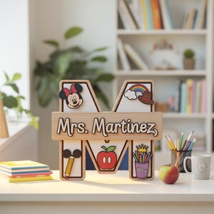 May include: A wooden letter "M" personalized with "Mrs. Martinez" and Disney-themed decorations. The design includes Minnie Mouse, a rainbow, an apple, and a pencil. The letter is on a desk with books and a mug.