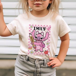 以下が含まれることがあります： クリーム色のTシャツ。ピンク色の漫画のキャラクターが鉛筆を持っています。Tシャツには「Emily」という名前がプリントされており、ピンクと白のチェック柄の背景があります。鉛筆には「First Grade」と書かれています。