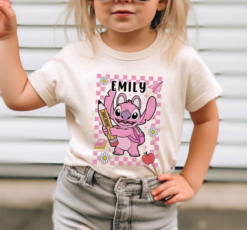 以下が含まれることがあります： クリーム色のTシャツ。ピンク色の漫画のキャラクターが鉛筆を持っています。Tシャツには「Emily」という名前がプリントされており、ピンクと白のチェック柄の背景があります。鉛筆には「First Grade」と書かれています。