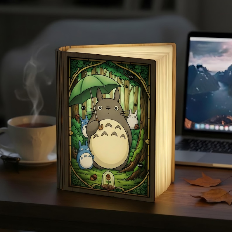 May include: A wooden book-shaped lamp with a colorful illustration of Totoro holding an umbrella in a forest scene. The lamp is open, emitting a warm glow, and is placed on a wooden surface. A cup of tea and a laptop are in the background.