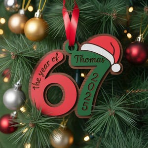 May include: A wooden Christmas ornament with the text "the year of 67 2025" and the name "Thomas". The ornament has a red and green color scheme with a Santa hat design. It hangs from a red ribbon.