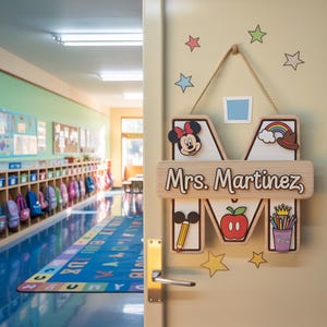 May include: Wooden classroom door sign in the shape of the letter "M" with the name "Mrs. Martinez". The sign features a Minnie Mouse head, a rainbow, an apple, and a pencil. The sign is hanging on a beige door.