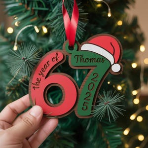 May include: A wooden Christmas ornament with the text "the year of 67" in red and "2025" in green. The ornament has a Santa hat and the name "Thomas". A red ribbon is attached for hanging.