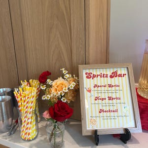 May include: A framed "Spritz Bar" sign with drink recipes, a vase of flowers, and a jar of yellow and red striped straws. The sign lists Aperol Spritz, Hugo Spritz, and Mocktail recipes. A clear pitcher of liquid is visible.