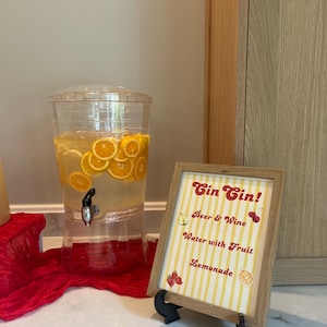 May include: A clear beverage dispenser filled with orange slices and liquid, next to a framed sign. The sign reads "Gin Gin!" with options for drinks like beer, wine, and lemonade. The sign has a yellow and white striped background.