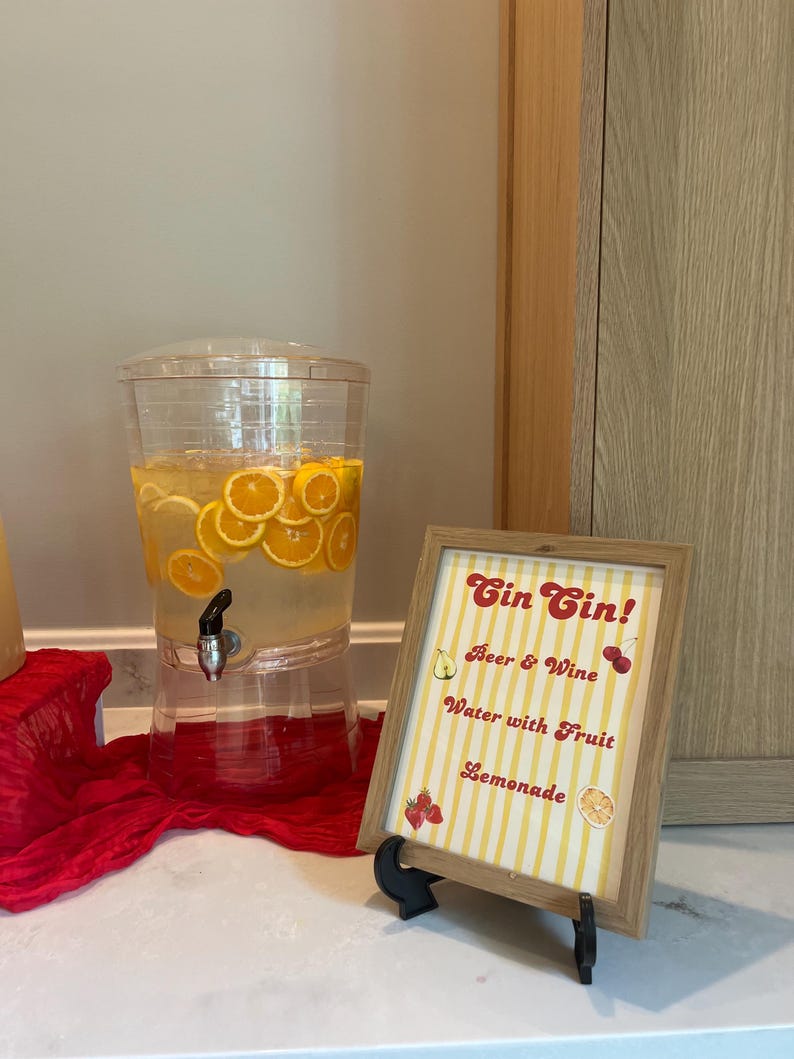 May include: A clear beverage dispenser filled with orange slices and liquid, next to a framed sign. The sign reads "Gin Gin!" with options for drinks like beer, wine, and lemonade. The sign has a yellow and white striped background.