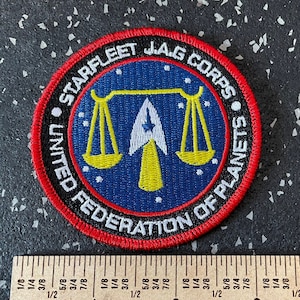 May include: A round patch with the text "STARFLEET J.A.G CORPS" and "UNITED FEDERATION OF PLANETS" in white lettering on a red border. The patch features a blue center with a white arrow and scales design. A ruler is next to the patch.